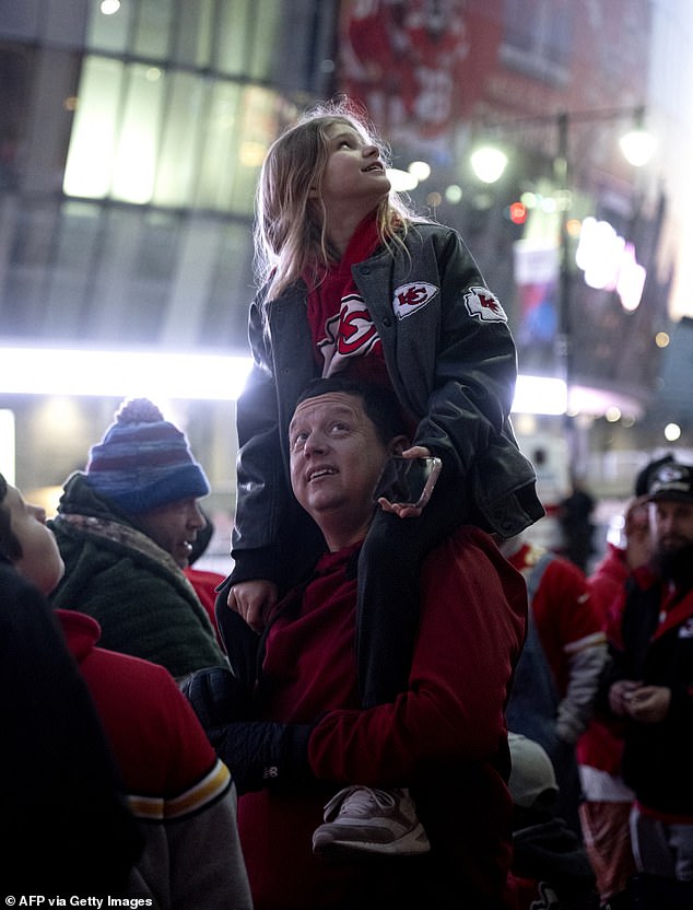 A young girl in a Chiefs sweater and jacket sits on her father's shoulders early in the morning