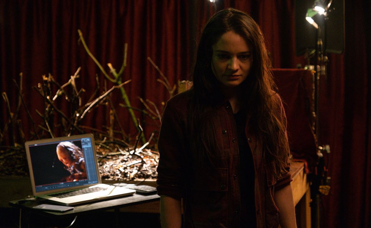 Stop-motion animator Ella (Aisling Franciosi) stands with her back to her set, a tangle of branches on a table in front of a red curtain, with a laptop open on the table, showing the warped, skull-faced girl from the film she makes in Stopmotion