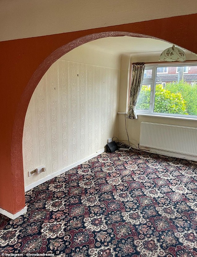 The room featured rust-red sofas, cream-colored walls, wooden furniture, a brass lampshade, a maroon and navy blue patterned carpet, and a tiled, coffee-colored fireplace.