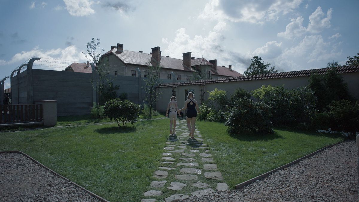 Two girls walk along a path in a walled garden with the guest houses of Auschwitz looming above them in The Zone of Interest