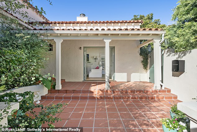 An outdoor paved terrace is framed by lush greenery and the driveway is hidden behind a double revolving wooden gate