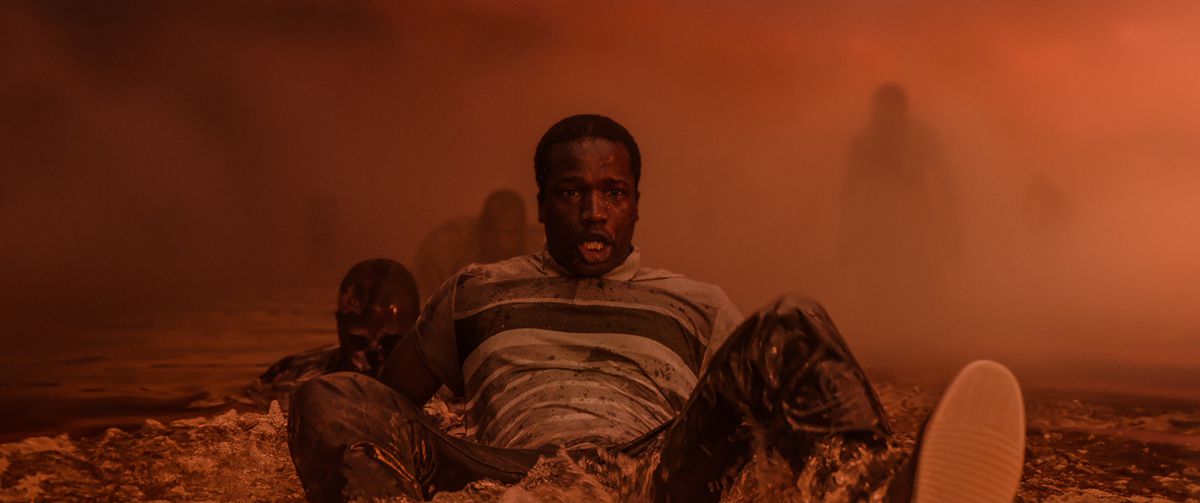 A terrified black man sits in a misty orange landscape, with menacing shadowy figures in the background