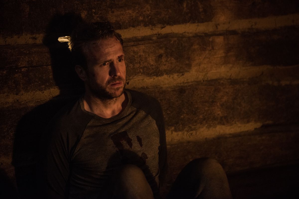 A man wearing a bloody shirt (Rafe Spall) sits immobilized against a brick wall lit by a nearby fireplace with a look of frustration on his face.