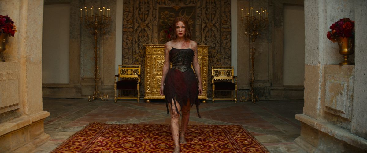 Elodie, played by Millie Bobby Brown, stalks the gilded palace corridors in a ruined, torn and burned dress, a look of determination on her face.