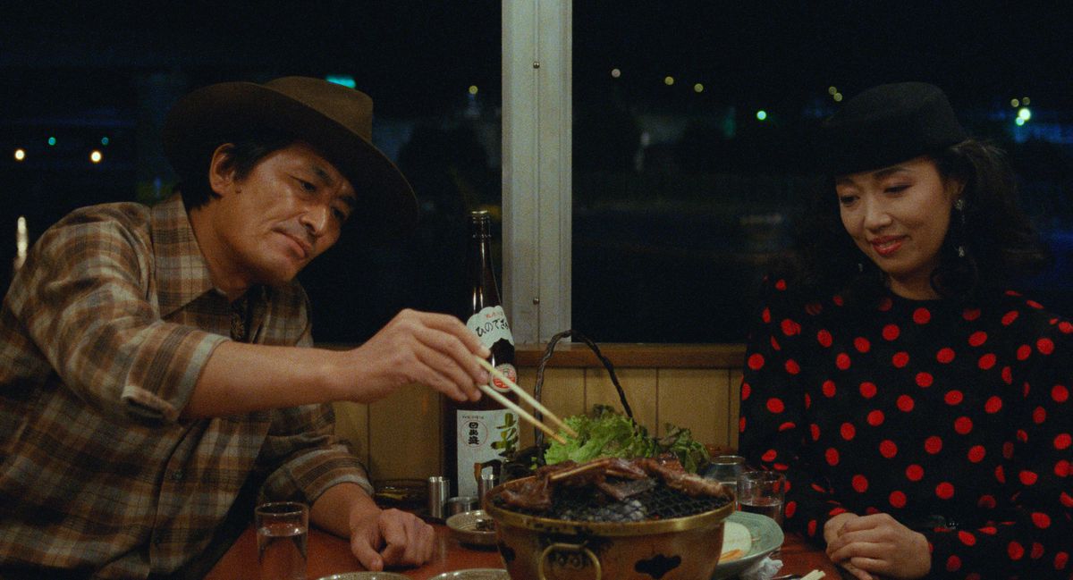 A man reaches with chopsticks for some meat on the grill in front of him, while a lady at the table smiles and watches