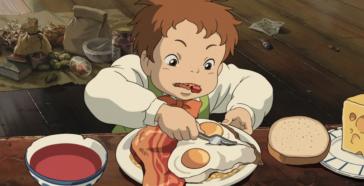 A young red-haired boy chews on a piece of thick bacon as he slices his lovingly detailed eggs sunny side up with a fork in a scene from Studio Ghibli's Howl's Moving Castle