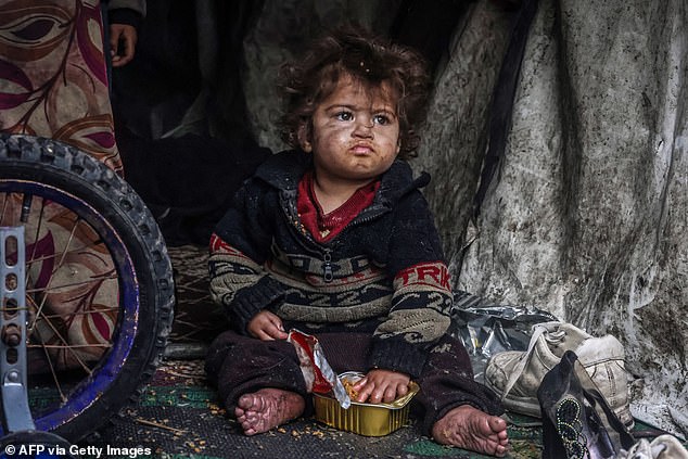 A displaced Palestinian child eats food from a box while sitting in a makeshift tent in a camp next to a street in Rafah on March 13, 2024