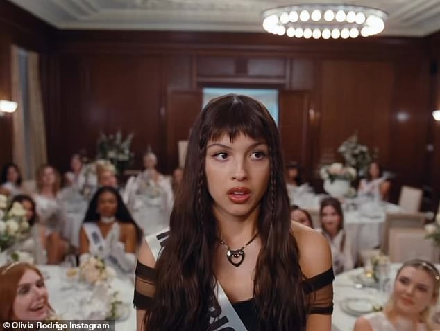 The teaser ends with Rodrigo, still dressed in black, facing forward towards the camera in front of the room full of ladies in white