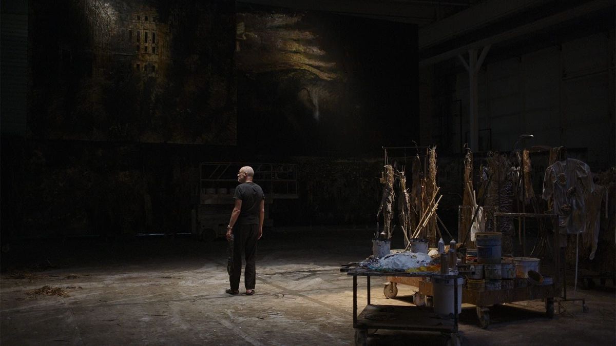 A man standing in a darkened studio, surrounded by art supplies and large paintings.