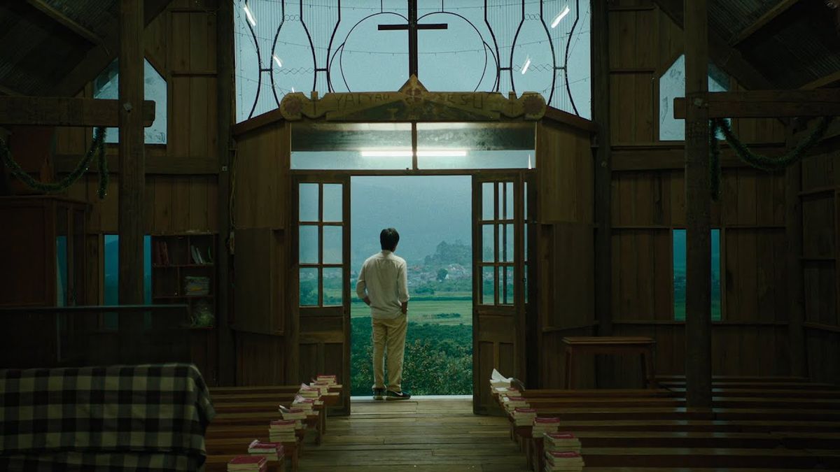 A man stands at the entrance of a church with a wide field of trees and grass in the distance.