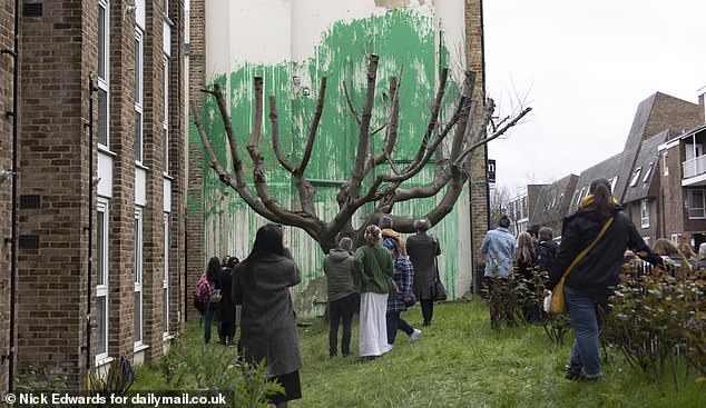 It is made with green paint behind a bare tree to resemble foliage, with a stencil of a person holding a pressure hose next to it.