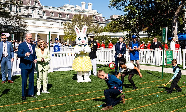 A life-size Easter Bunny stands next to President Joe Biden and First Lady Jill Biden, who are shown smiling as they watch the children and participate in the Easter scroll