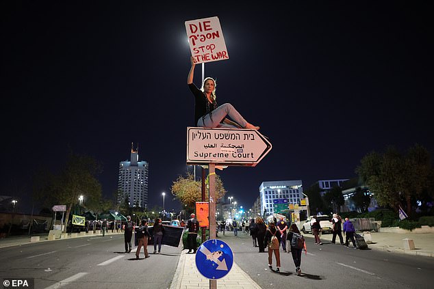 A protester sits atop a sign pointing to the Israeli Supreme Court, holding a sign that reads 