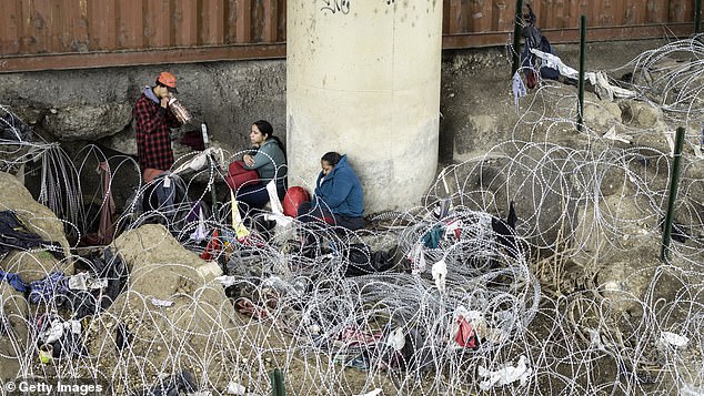 In an aerial view, immigrants wait under an international bridge after crossing the Rio Grande from Mexico and passing through coils of barbed wire on March 17, 2024 in Eagle Pass, Texas