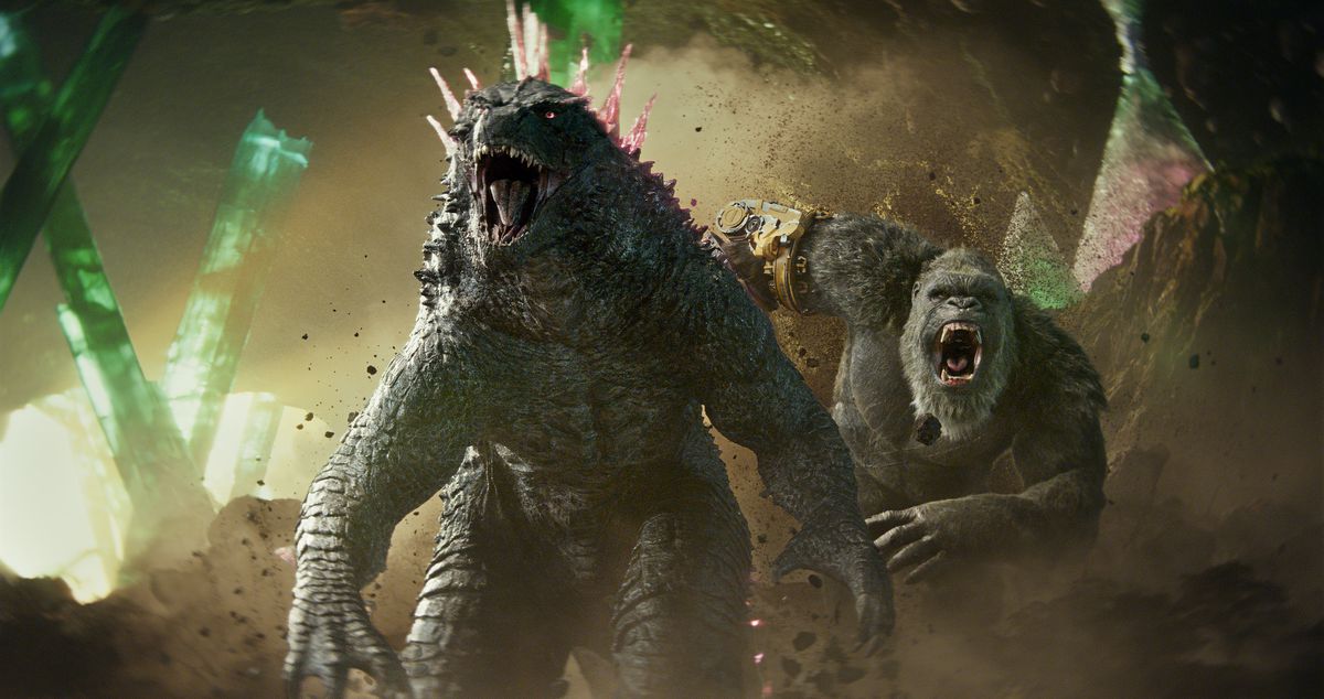 Godzilla, now with pink spikes on his back, roars as King Kong, now with a robotic hand, roars behind him as they stand together in a cave in Godzilla x Kong: The New Empire