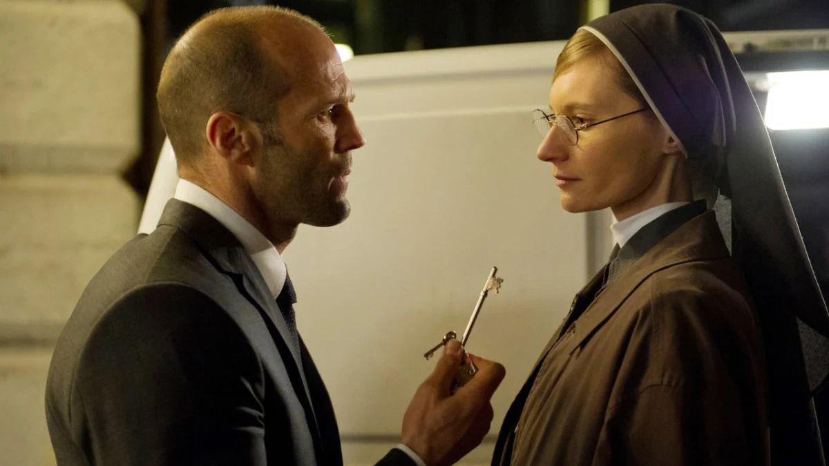 A bald man in a suit holds up a pair of keys to a woman with glasses in a nun suit.