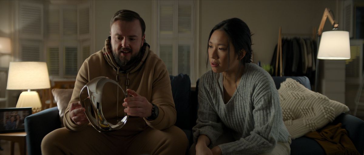 John Bradley as Jack Rooney and Jess Hong as Jin Cheng sit on a couch and look at the chrome VR headset in episode 102 of 3 Body Problem