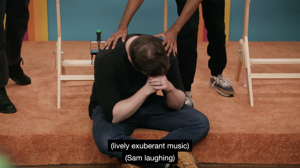 A screencap from Game Changer's 'Bingo', showing Brennan Lee Mulligan sitting on the floor with his head in his hands.  The subtitles read: “(lively, exuberant music)” and “(Sam laughs).”