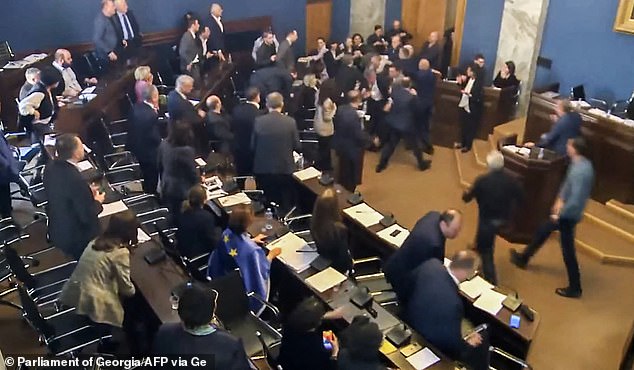 As Elisashvili and the speaking politician fall off the stage together, a mass of bodies surround them and try to subdue the MP.