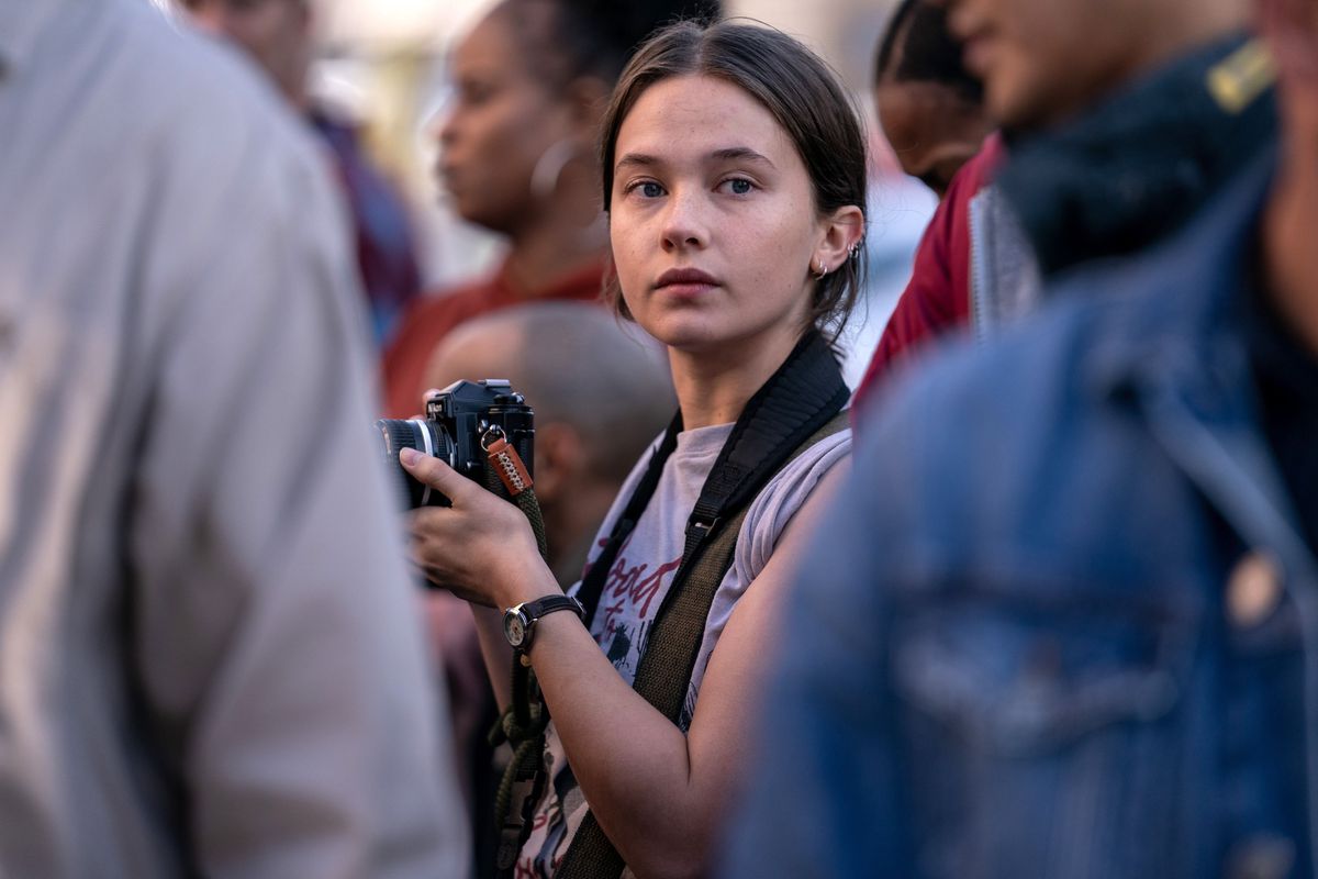 Jessie (Cailee Spaeny), a young photojournalist holding a camera, stands in the middle of a crowd in Alex Garland's Civil War