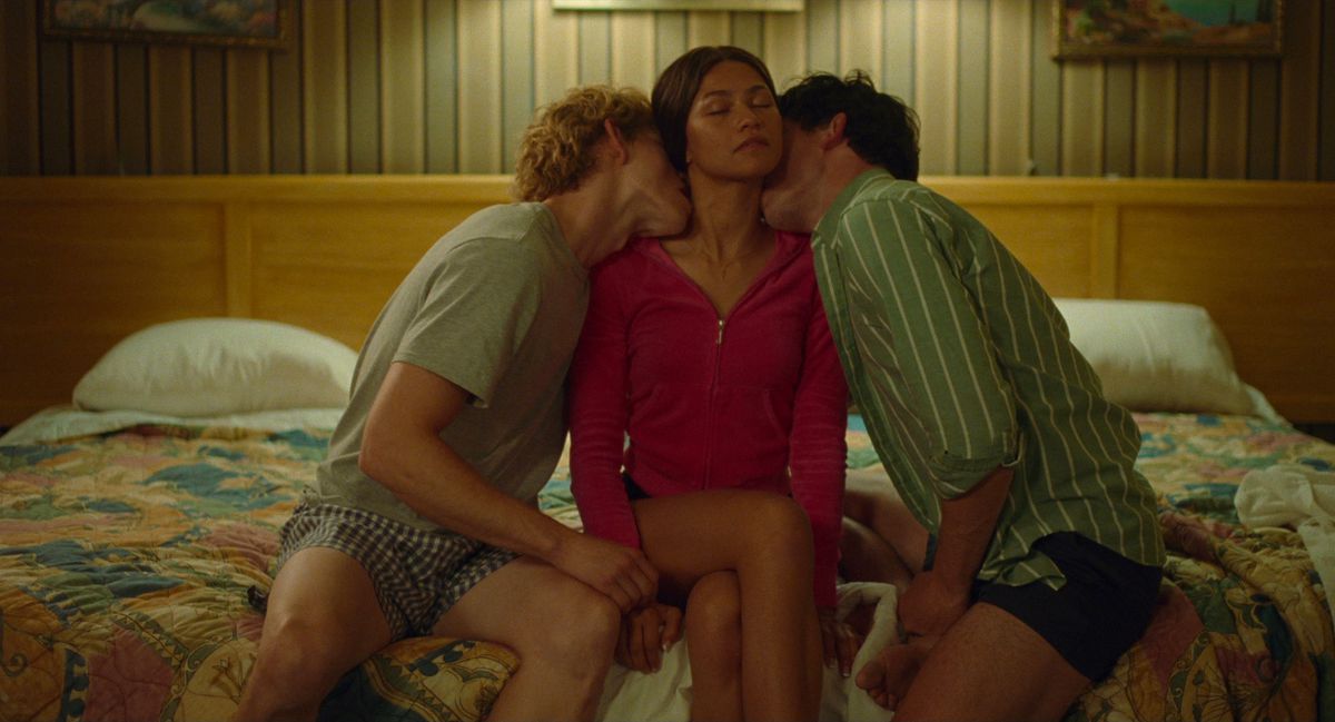 Teenager Tashi (Zendaya, in a bright red sweater) sits on a hotel bed, eyes closed, chin up, hands at her sides, while Art (Mike Faist) and Patrick (Josh O'Connor) sit on either side of her, each passionately holding her neck kissing, in Challengers