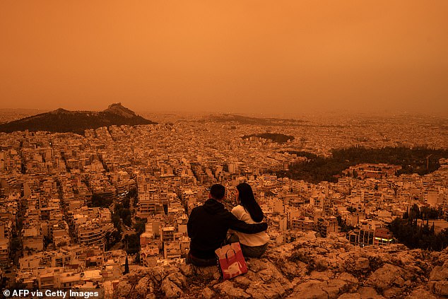 A couple pictured sitting on the hill overlooking a Martian Athens after southerly winds painted the city in an unusual hue