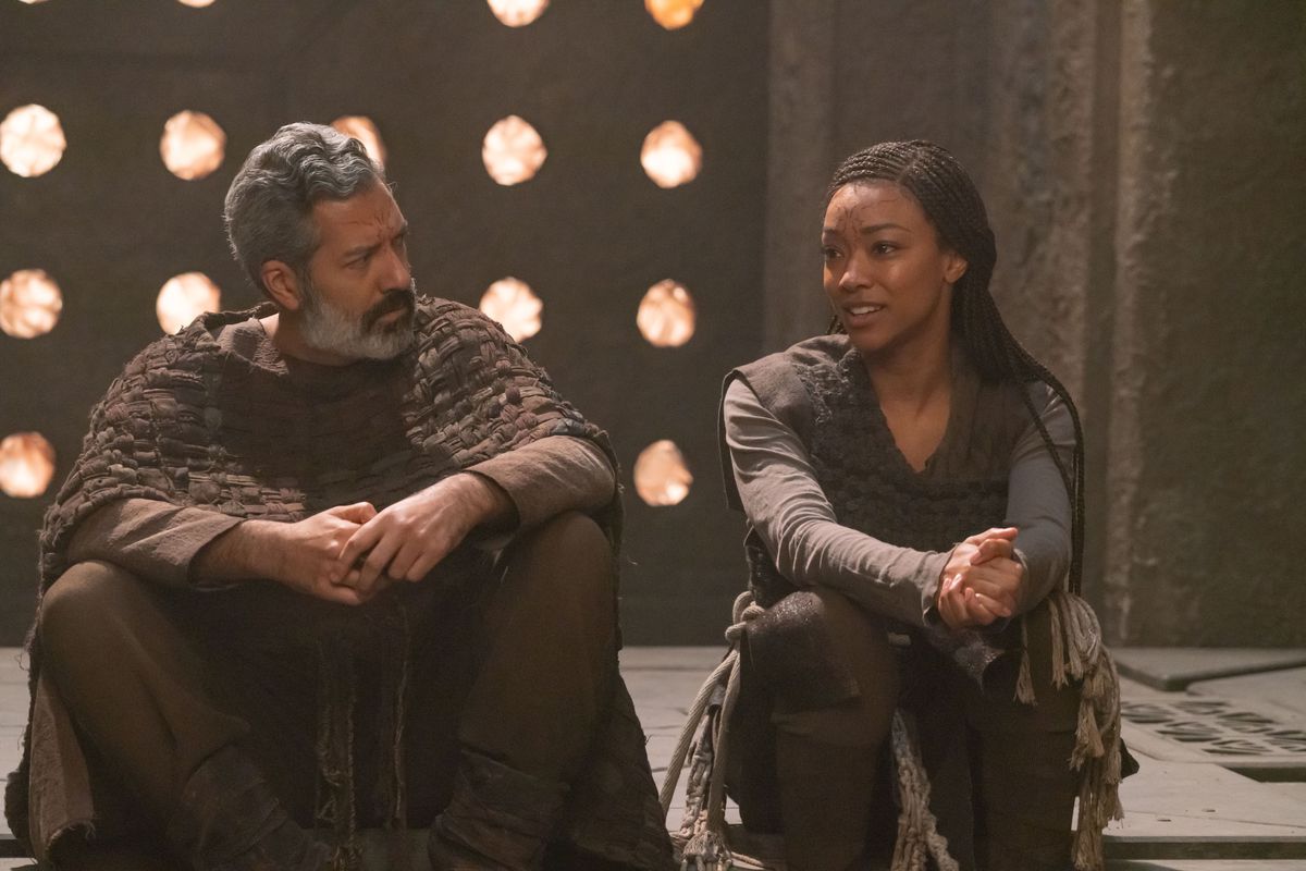 L-R Alfredo Narciso as Ohvahz and Sonequa Martin-Green as Michael Burnham in Star Trek: Discovery.  They wear handmade alien garments and talk quietly while sitting on the floor in a stone room.