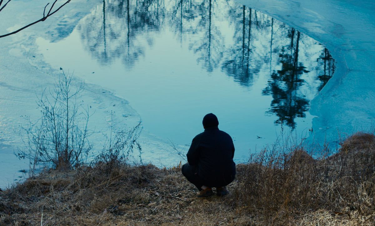 A figure in a coat and hat, crouched from behind at the edge of a half-frozen lake as the forest reflects on its surface in the film Evil Does Not Exist.
