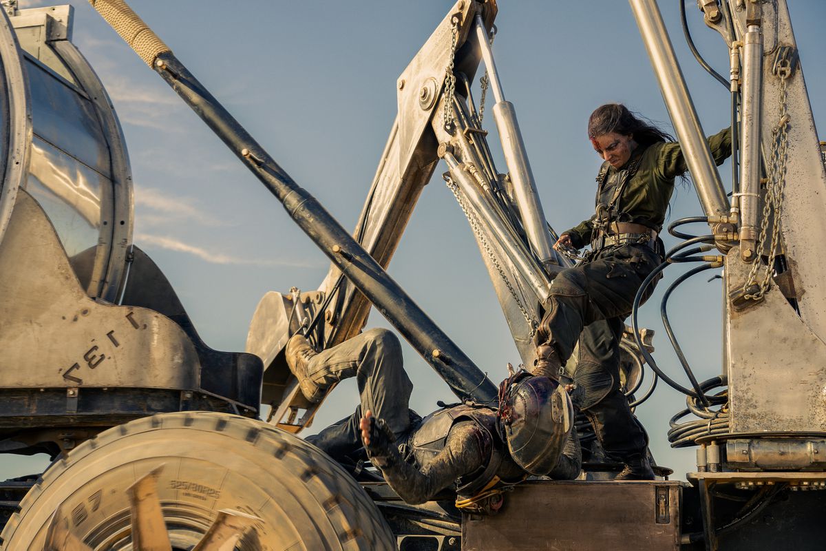 Furiosa (Anya Taylor-Joy), a leather-clad warrior with long braided hair, stands on the back of a steam shovel and kicks a helmeted, prone opponent in the face, in George Miller's Furiosa