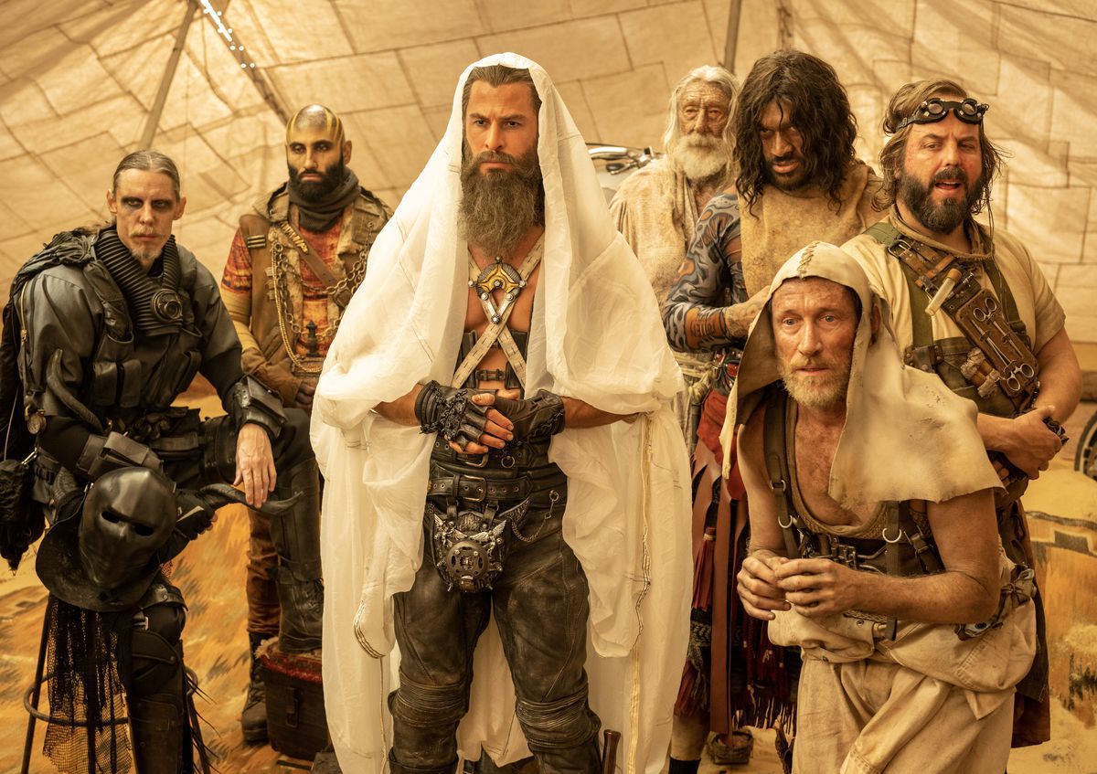 Dementus (Chris Hemsworth), the bearded leader of a group of Wasteland warriors, stands in a tent with a clean white cloth draped over his head, reaching down to his feet, surrounded by male acolytes of various ages in George Miller's Furiosa