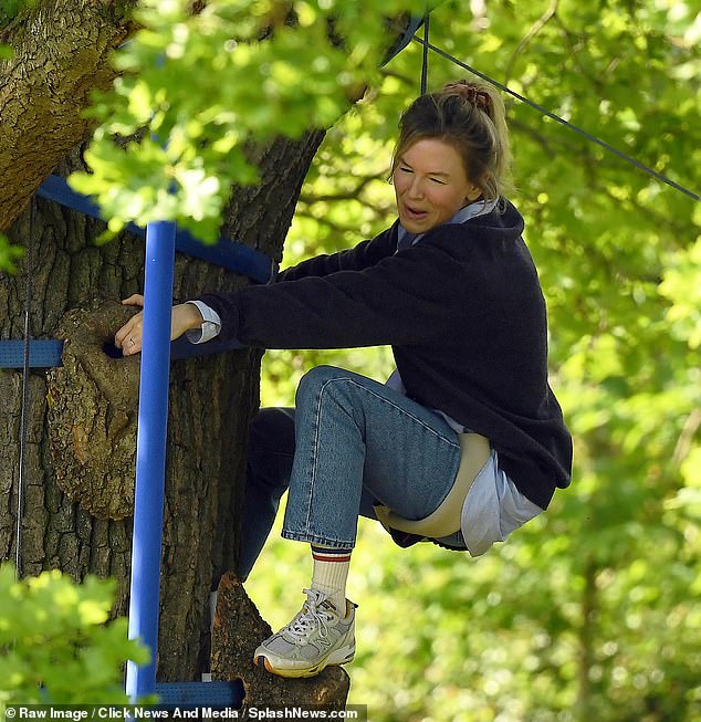 A smiling Mrs. Zellweger hugs the tree and wears jeans with a shirt and sweater for the scene