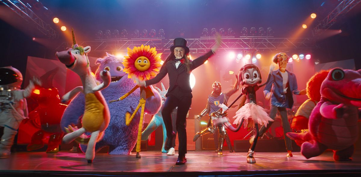 Bea (Cailey Fleming), in a suit and top hat, dances on a stage under bright lights, surrounded by CG creations representing children's imaginary friends: a bipedal, big-nosed unicorn in a yellow suit, an astronaut, a sunflower with a face and legs, a large purple furry creature, and more