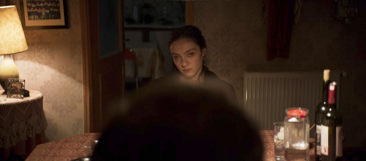 A woman sits at a dining table, dimly lit.  She looks vaguely angry.  We look at her over someone else's head.