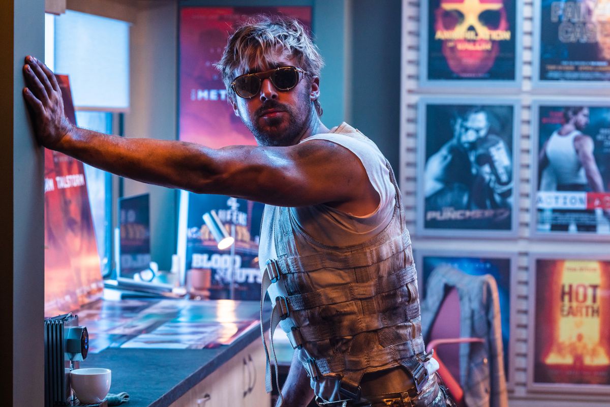 In a scene from The Fall Guy, stuntman Colt Seavers (Ryan Gosling, wearing sunglasses, a scruffy sleeveless vest and a scowl) leans with one arm against a wall in a room full of neon-colored movie posters.