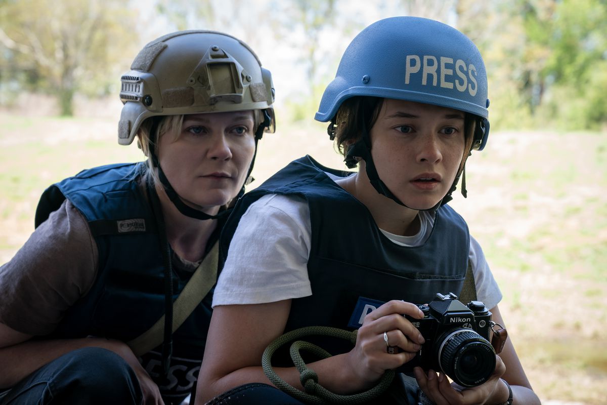 Photojournalists Lee Miller (Kirsten Dunst) and Jessie (Cailee Spaeny) huddle together, Jessie holding her camera and wearing a bright blue helmet that reads 