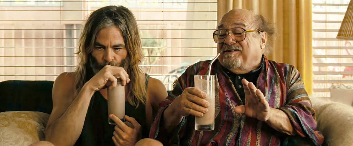 A long-haired and disheveled Chris Pine thoughtfully sips a milkshake as Danny DeVito (also holding a milkshake) explains something to him