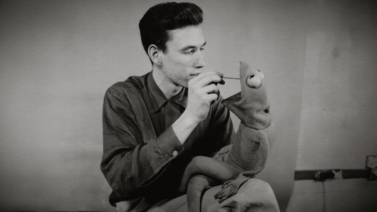 A black and white image of a man adjusting something in the mouth of a Kermit the Frog doll 