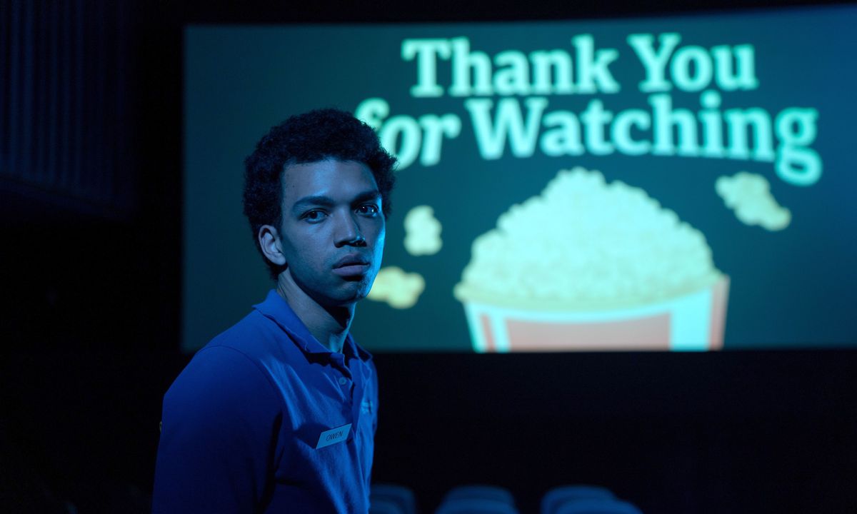 Twenty movie theater worker Owen (Justice Smith) stands in a dark movie theater looking at the camera, with a slide on the screen behind him that reads 