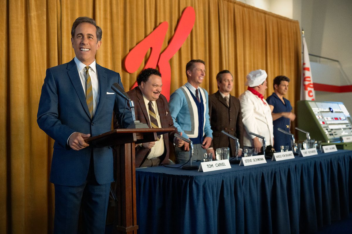 Seinfeld stands behind a podium in front of the Kellogg's K alongside a panel of other people in the Netflix film Unfrosted.