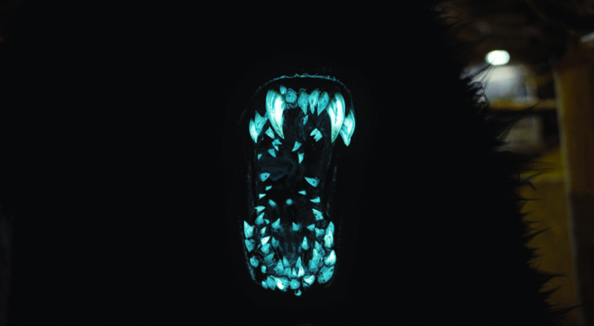A mouth filled with blue glowing teeth, placed in the jaw of a jet black hairy monster.