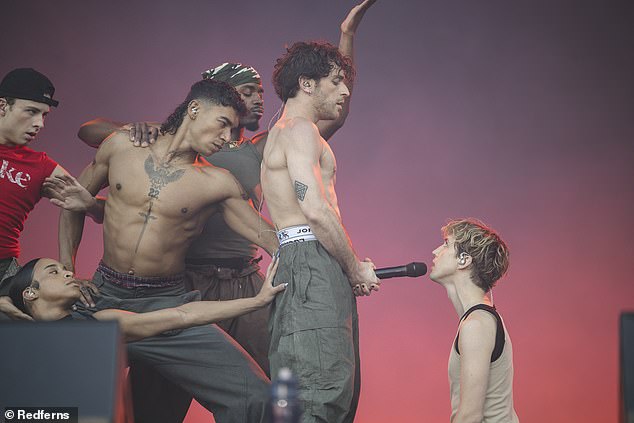 Troye kneels on the floor while one of his dancers holds the microphone in an X-rated position.  Troye sings, “Boy, can I be honest?”  before taking the microphone back