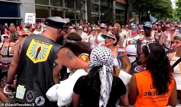 A large man in a leather vest and cap stopped a large group of women in colorful outfits playing drums as they tried to continue their parade route