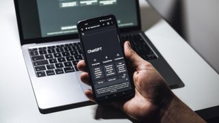 A hand holding a phone running ChatGPT, in front of a laptop