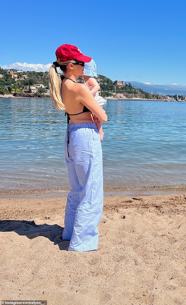 Cricketer Nathan Lyon's wife, wearing a black bikini top, long blue pinstripe pants and a red baseball cap, posed with her baby on the shore of a bay