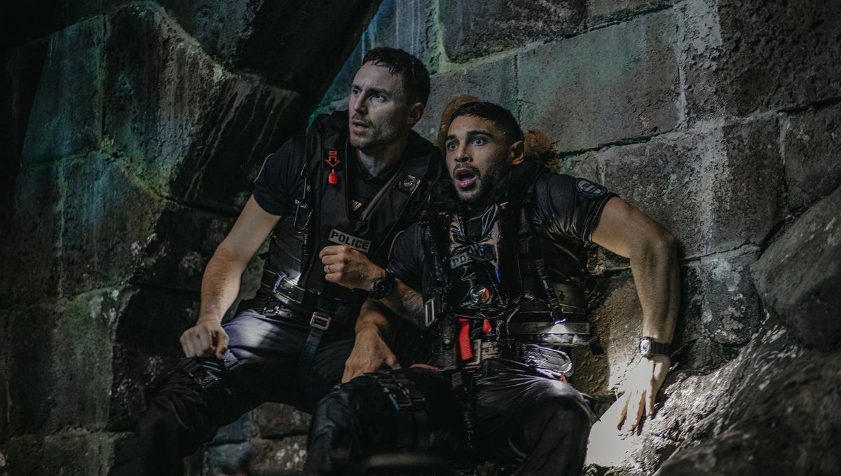 Police Sergeant Adil (Nassim Lyes) and a fellow officer, both soaking wet and in tactical gear, press themselves against a stone wall in an underground Parisian water reservoir in Xavier Gens' Netflix shark thriller Under Paris