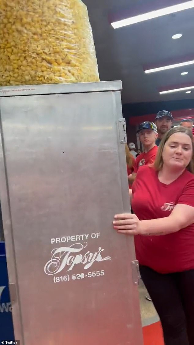 A woman dressed in red with pursed lips pushing a large metal container precariously holding a bag of popcorn