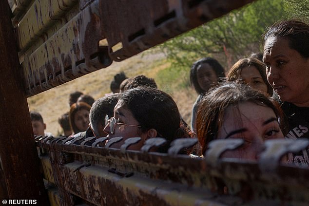 Migrants from South and Central America look through a hole in the border wall at the United States before crossing Boulevard, California while lining up to enter the United States from Tecate, Mexico