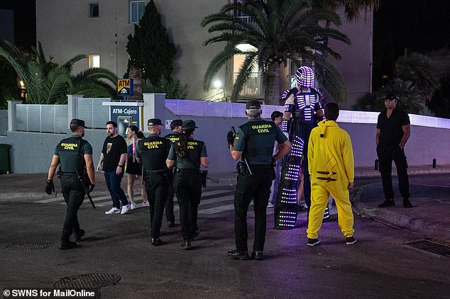 A group of police officers patrol the streets of Magaluf, while a man in an elaborate costume, who appears to be walking on stilts, walks in the opposite direction