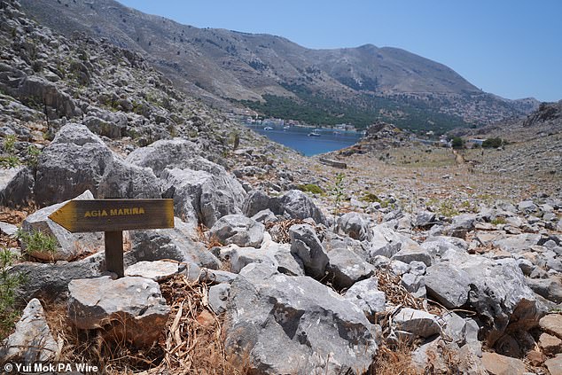 A directional sign pointing towards Agia Marina on a rocky path in the hills of Pedi (Pedi center pictured in the distance, right), a small fishing village