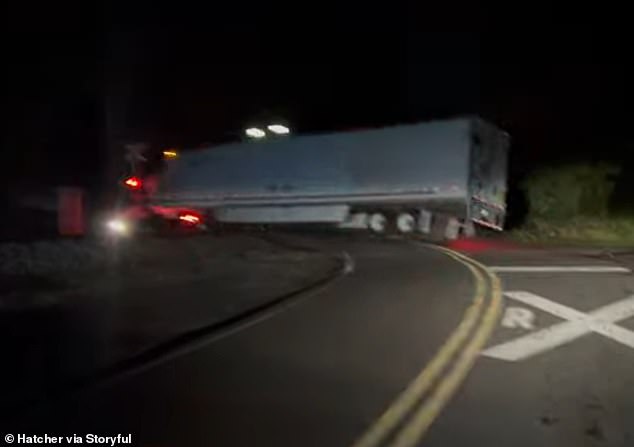 The footage shows the trapped semi-truck stretching across the train tracks.  It is night and the railway crossing signal is flashing red